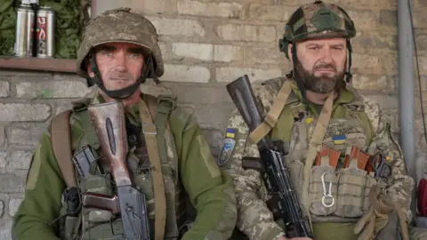 Two Ukrainian soldiers wearing helmets and carrying guns look   into the camera lens