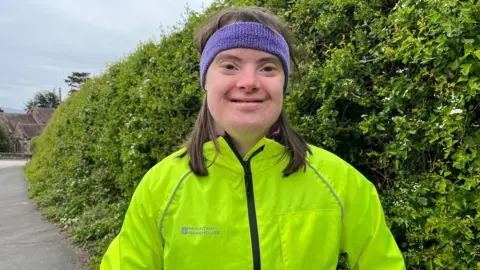 Serena has brown hair and a purple sweatband on her head. She is wearing a bright yellow windbreaker and is smiling. She is standing in front of a green bush on a path.
