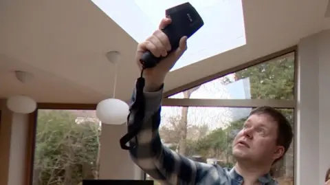 A man in a plaid shirt and a woman look upward inside a modern house with large windows. The man is holding a thermal imaging device up toward a skylight.