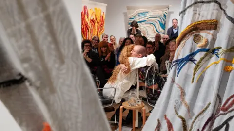 London Gallery Weekend/Linda Nylind Two people embracing with a crowd around them clapping. They are surrounded by long, colourful tapestries.
