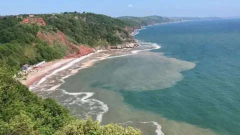 Oddicombe Beach in Torquay - blue sky and sea. Red rockface and green, vegetation covered cliffs
