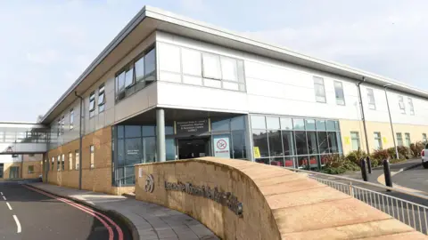 ELHT The exterior of a two-storey building, with yellow brick on the ground-floor walls and white cladding the first-floor walls. 
