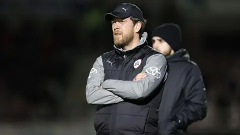 Getty Images Manager Darrell Clarke is standing with his arms crossed on a pitch. He is wearing a black gilet with grey sleeves visible underneath and a black cap.