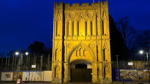 An illuminated ornate cream stone tower building, with hoardings either side.