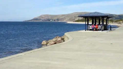 Stephen McKay/Geograph Two people sitting in a hut on a concrete promenade in front of water with hills in the background
