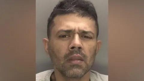 A custody image of Harinderpal Athwal, a man with a short black beard and moustache and short hair is looking at the camera with a slight frown on his face