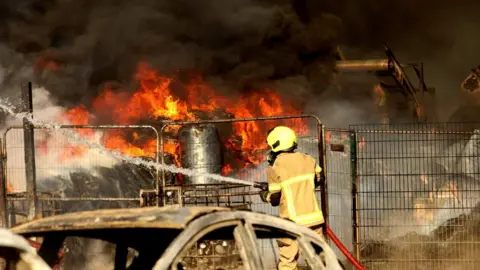 A firefighter tackling a large fire behind a chain-link fence. In the foreground, burnt out cars can be seen.