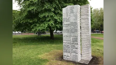 The Cambridge Rules statue on grass on the edge of Parker's Piece, surrounded by trees. It is a tall grey stone sculpture that has the football rules on it in many languages.