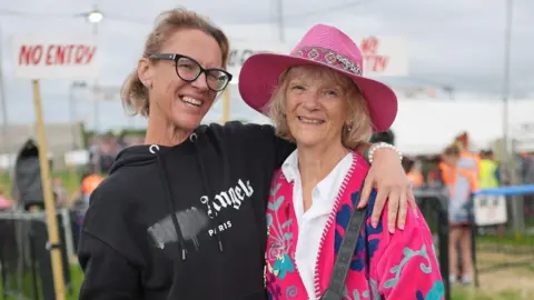Two women stand side by side in front of the gates at Glastonbury Festival. The daughter, who is wearing a black hoodie, has her arm around her mum, who is wearing a pink sunhat and a pink, patterned jacket. They are both smiling. Behind them stewards can be seen allowing people into the site.