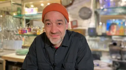 Javier Tanke is stood behind the bar, wearing an orange beanie hat and dark shirt. He has a short, grey beard.