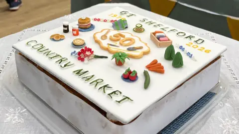 Tom Jackson/BBC A square cake is sat on a table which has a white patterned table cloth over it. The cake has GT SHELFORD COUNTRY MARKET written on it in green icing and has different items made out of icing on it, including carrots, cucumber and flowers.