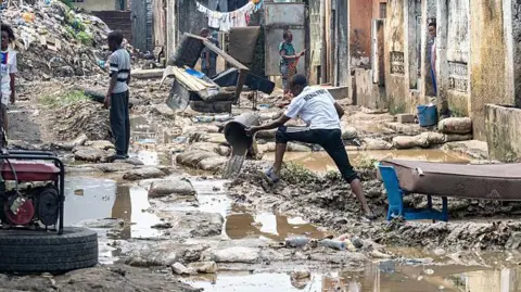  A resident clears water with a bucket amid debris in a residential area affected by flooding following torrential rains in Kinshasa, on 15 June  2025