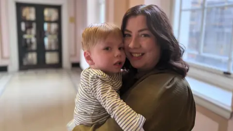 Mary Frances Loughran holding her son Finn. She has shoulder-length brown hair, wearing a khaki green top. Finn Has blonde hair and wearing a green and white striped top.