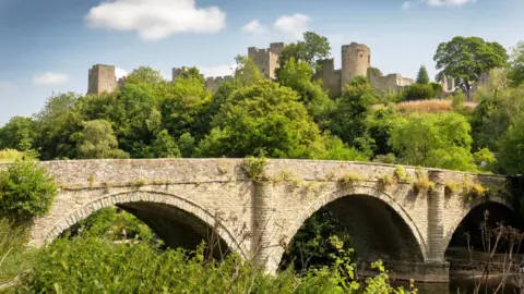 A brick bridge over a river with three arches. Around it are green bushes and trees. In the background above is Ludlow Caste atop a hill