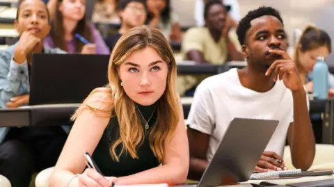 Getty A female student sits on the front row of a lecture hall. She has long light brown hair and wears a black vest top witha. silver necklace. She is looking at the lecturer while writing with a black pen on a notepad. Her laptop is open in front of her.