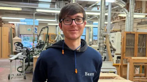 A young man wearing a blue sweatshirt with a black hoodie underneath. He has brown hair and glasses and is smiling. He is standing in front of a work shop which has multiple wooden projects and machinery. 