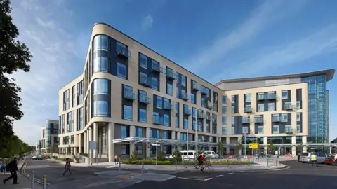An external shot of the main building at Southmead Hospital. The structure is cream, with large windows and some balconies. In the foreground is a parking area with a short glass roof over it