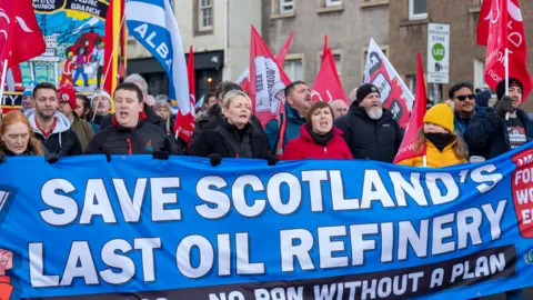 A group of protestors carrying flags and chanting slogans march along the road. Five protestors at the front are holding a large blue banner which reads: "Save Scotland's last oil refinery. Oil and gas - no ban without a plan". 