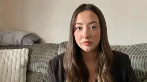 Laura is a young woman with long, brown hair. She is staring at the camera. She is sitting on a grey sofa and is wearing a brown pinstripe blazer and a black t-shirt.