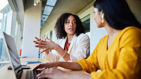 Getty Images Two women working together in an office, with one gesturing with her hands as she speaks, and the other typing on a keyboard