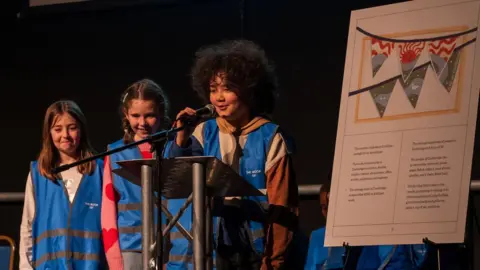 Liam Togher Three young girls wearing blue hi-vis vests standing on stage, next to a large version of their book. One of the girls is standing behind a lectern and speaking into a microphone. The other two look excited but have nervous smiles.