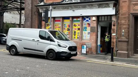A police officer stands guard in the doorway of a shop. A grey van is parked outside