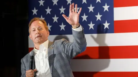 Texas Attorney General Ken Paxton waves while leaning and holding a microphone in front a US flag backdrop.