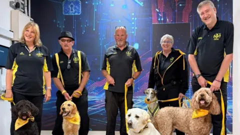 Leonardo Five different breeds of dog, including a golden retriever and labrador, are wearing yellow neckerchiefs and are being held on leads by charity workers wearing black and yellow polo tops.