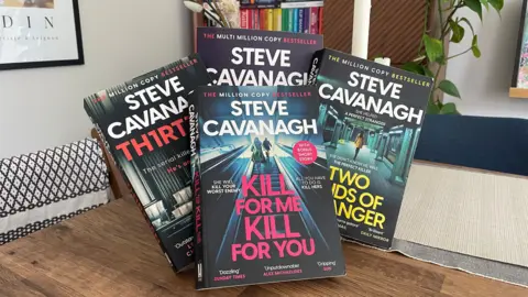 A handful of books from Steve Cavanagh sitting on a table in front of a book case.