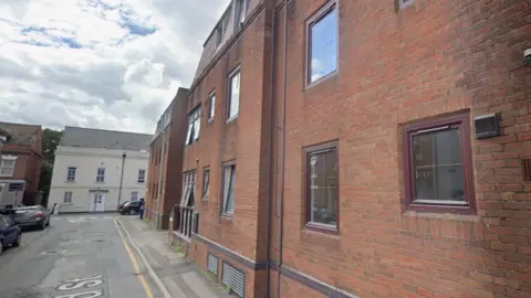 Google An aged block of flats of about three storeys on a side road in a city centre. The building is of brown brick and brown windows. Cars are parked on the road leading up to a T-junction, where there are terraced houses. It is a mostly cloudy day with a small patch of blue sky.