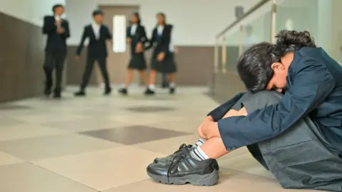 Getty Images A girl wearing school uniform sits on the floor in a school corridor with her knees drawn up and her head resting on them. Out of focus in the background are four other children in uniform. 