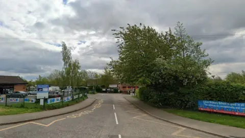 Google The entrance to a Upton Primary School. There are colourful posters on the approach to the school with trees and greenery surrounding the site. 