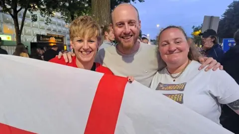 Two women hold an England fan and smile at the camera with a man smiling in the middle
