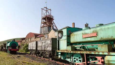 A view of Big Pit in Blaenavon - ageing, rusting green trains can be seen with wagons attached to the back of them on a disused railway track with grass growing between the rails. Industrial buildings of an old coal mine can be seen behind them and a hillside is in the distance 
