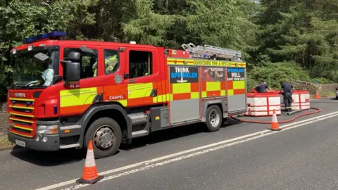 Derbyshire Fire and Rescue Service A fire engine parked on the side of the road next to woodland while crew members use a water carrier to help pump water into the wooded area. 