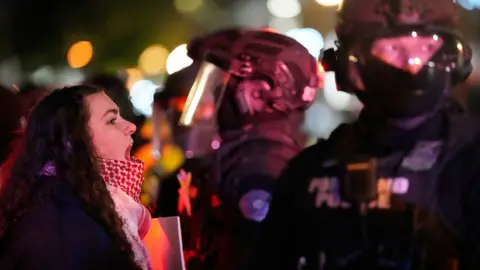 AP A young woman who seems to be shouting confronts an armed member of law enforcement, who is wearing a helmet