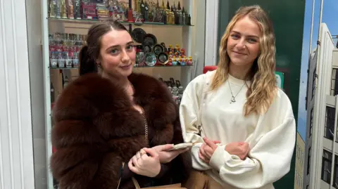 Taylor is wearing a brown fluffy coat and Lucy is wearing a beige sweater as they stand in front of a shop window displaying tourist trinkets.