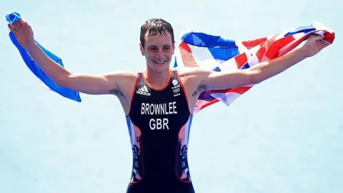 Alistair Brownlee celebrating his gold medal win at the Rio De Janeiro Olympic games in 2016. He is wearing a black Team GB running vest with BROWNLEE GBR written on the chest and black shorts. His arms are outstretched and he is holding the Union Jack flag behind his back, arms and shoulders.