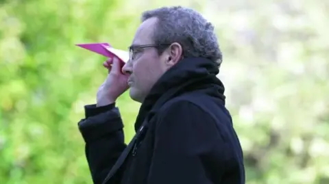 a man in a black coat wearing glasses holds a pink paper aeroplane, ready to fly it into the air