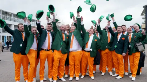 Men dressed in orange trousers, with green jackets. They have orange ties and pocket squares. All of them are smiling and throwing their green hats in the air.