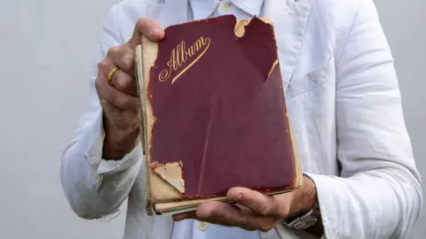 Hansons Auctioneers A book with a battered red hardback cover, with the word Album written in gold lettering, held in a pair of hands