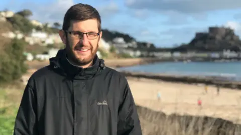 A man with dark hair and beard, wearing glasses and a black coat. He is standing in the foreground with a beach scene out of focus behind him.