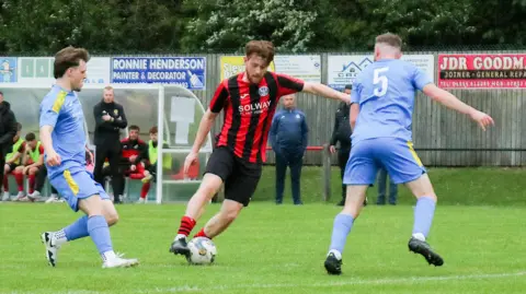 David McDonald A footballer in a red and black striped top and black shorts has the ball at his feet while two players in blue kit with yellow shoulder stripes close in on him