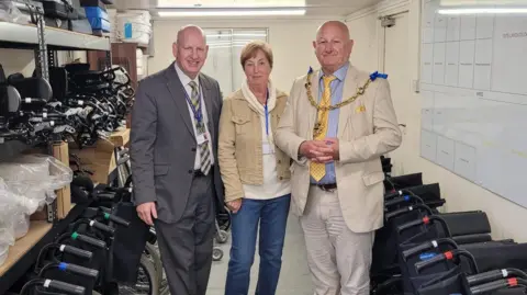 Two bald men in suits standing either side of a woman in a white hoodie and beige denim jacket. There are wheelchairs parked either side of the group.