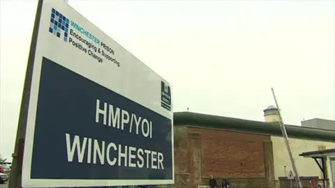 A sign with the words HMP/YOI Winchester in white text on a blue background, in front of the large brick perimeter wall of the prison.