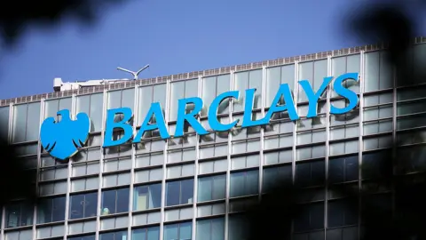 The Barclays logo is seen on the side of the Barclays building in Canary Wharf, with foliage silhouetted out of focus in the foreground.