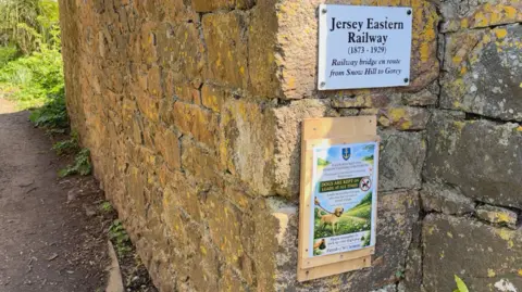 A stone wall beside a narrow path displays two signs: one commemorating the Jersey Eastern Railway bridge and another advising that dogs must be kept on leads, with greenery growing along the wall and path