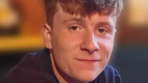 A young man, Henry Farron smiles. He has gingery hair and is wearing a navy jumper. The background in the image is blurred.