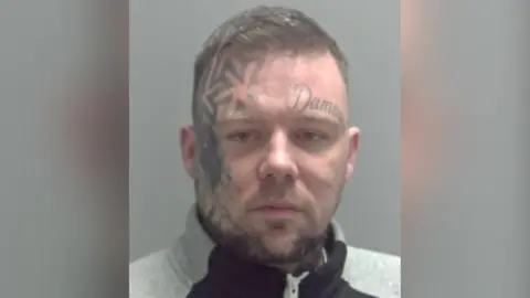 Suffolk Police Daryl Taylor looks directly at the camera in a custody mugshot, he has a tattoo on his face and is wearing a grey jumper.