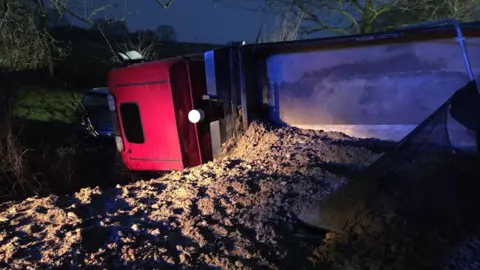 A lorry is pictured at night. It appears to be overturned with its contents spilling on to the road.